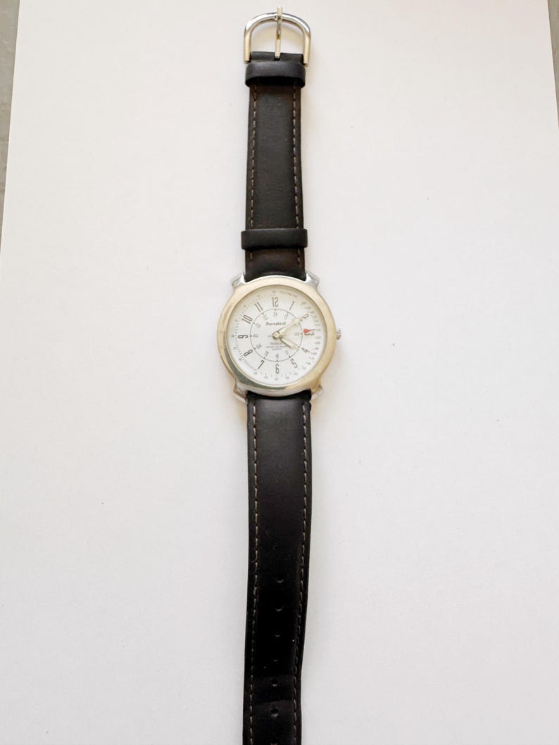 May include: A silver-toned wristwatch with a white face and black leather band. The watch has a round face with black numerals and hands. The black leather band is stitched and has a silver buckle.