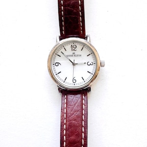 K&ouml;nnte beinhalten: Anne Klein Armbanduhr mit wei&szlig;em Zifferblatt, silberfarbener L&uuml;nette und braunem Lederarmband. Das Zifferblatt zeigt das Anne Klein Logo, schwarze Ziffern und Zeiger. Das Armband ist mit wei&szlig;em Faden gen&auml;ht.