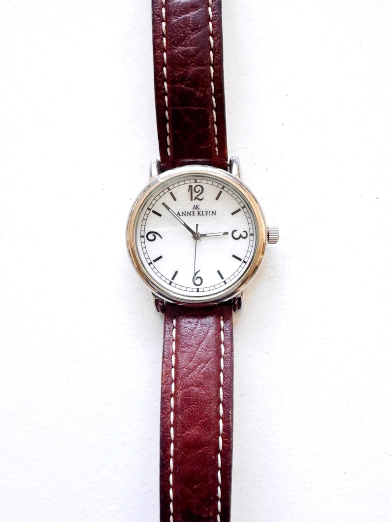 K&ouml;nnte beinhalten: Anne Klein Armbanduhr mit wei&szlig;em Zifferblatt, silberfarbener L&uuml;nette und braunem Lederarmband. Das Zifferblatt zeigt das Anne Klein Logo, schwarze Ziffern und Zeiger. Das Armband ist mit wei&szlig;em Faden gen&auml;ht.