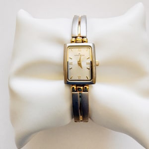 May include: A rectangular Anne Klein wristwatch featuring a two-tone silver and gold metal band and case. The watch face is light-colored with gold-tone hour markers, including Roman numerals at the twelve and six o'clock positions, and gold hands.