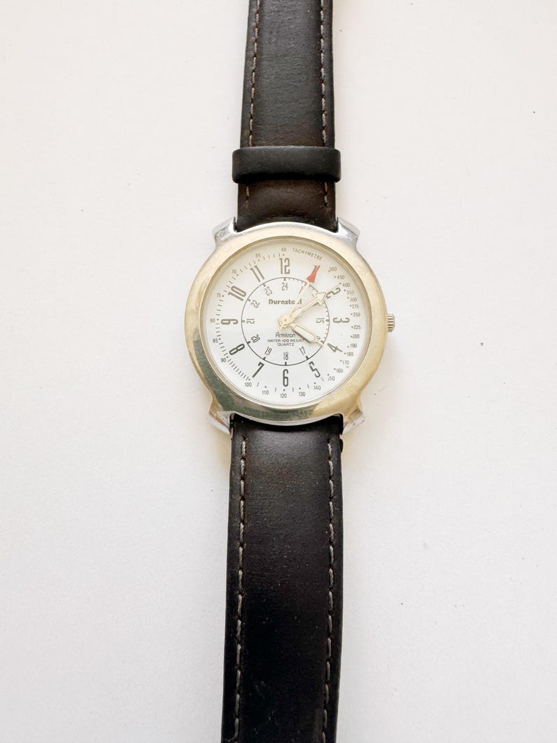 May include: A wristwatch with a white face, silver-colored bezel, and a dark brown leather band. The watch face displays the brand name "Burnsto A" and has black numerals and hands. The band is stitched with a lighter thread.