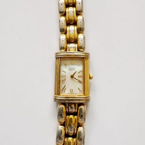 May include: A rectangular Citizen Elegance wristwatch with a white face, gold-tone Roman numeral hour markers, and gold-tone hands. The watch features a two-tone gold and silver metal link bracelet with a polished finish and a side adjustment crown.