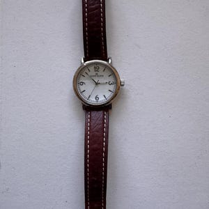 K&ouml;nnte beinhalten: Eine silberfarbene Anne Klein-Uhr mit wei&szlig;em Zifferblatt und schwarzen Ziffern. Sie hat ein braunes Lederarmband mit wei&szlig;en N&auml;hten. Das Zifferblatt zeigt das Anne Klein-Logo und die Zahlen 3, 6, 9 und 12.