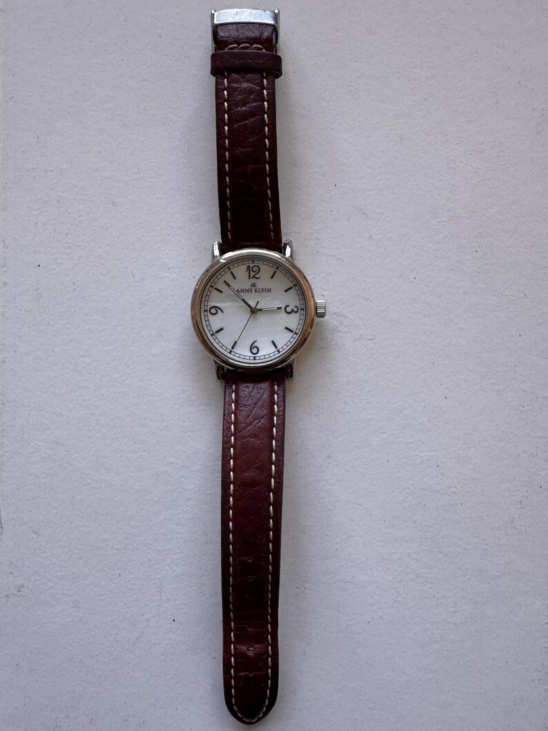 K&ouml;nnte beinhalten: Eine silberfarbene Anne Klein-Uhr mit wei&szlig;em Zifferblatt und schwarzen Ziffern. Sie hat ein braunes Lederarmband mit wei&szlig;en N&auml;hten. Das Zifferblatt zeigt das Anne Klein-Logo und die Zahlen 3, 6, 9 und 12.