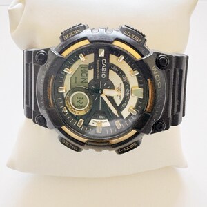 May include: Black Casio digital watch with gold accents, displayed on a white pillow. The watch face shows the day, date, and time. Features include a timer, search function, and water resistance. The watch has multiple buttons for adjusting settings.