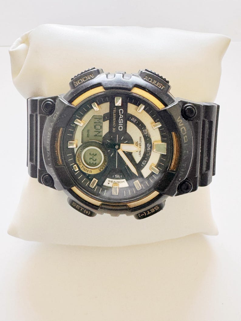 May include: Black Casio digital watch with gold accents, displayed on a white pillow. The watch face shows the day, date, and time. Features include a timer, search function, and water resistance. The watch has multiple buttons for adjusting settings.