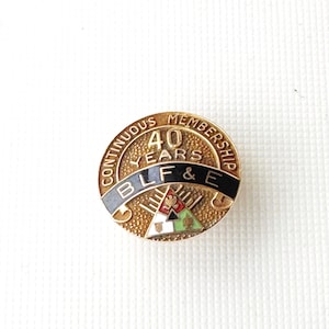 Puede incluir: Pin dorado con el texto "40 YEARS" y "CONTINUOUS MEMBERSHIP". El pin también presenta las letras "BLF & E" y un pequeño emblema con detalles rojos, verdes y blancos. Forma redonda.
