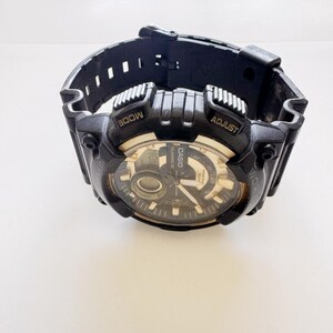 May include: Black Casio digital watch with a black band. The watch face is black and gold with the Casio logo. The watch has the words "MODE" and "ADJUST" on the top. The watch is on a white surface.