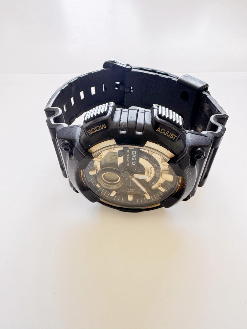 May include: Black Casio digital watch with a black band. The watch face is black and gold with the Casio logo. The watch has the words "MODE" and "ADJUST" on the top. The watch is on a white surface.