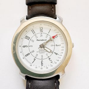 May include: A silver-toned Armitron Durasteel quartz watch with a white face, black numerals, and a tachymeter scale. The watch has a black leather band. The watch face displays the text "Armitron Water 100 Resist Quartz".