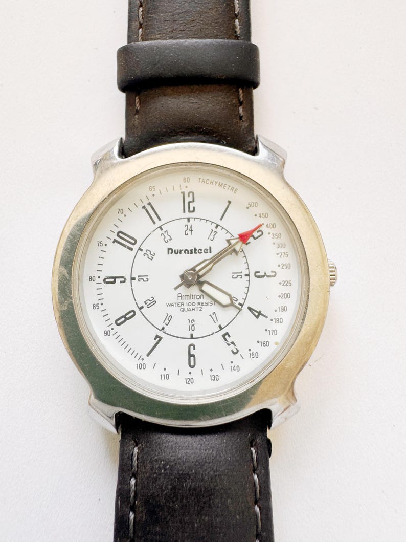 May include: A silver-toned Armitron Durasteel quartz watch with a white face, black numerals, and a tachymeter scale. The watch has a black leather band. The watch face displays the text "Armitron Water 100 Resist Quartz".