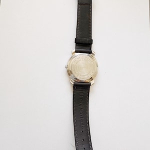 May include: A wristwatch with a black leather band and a silver-colored metal case. The watch band is connected to the case with a silver buckle. The back of the watch case has text and a logo.