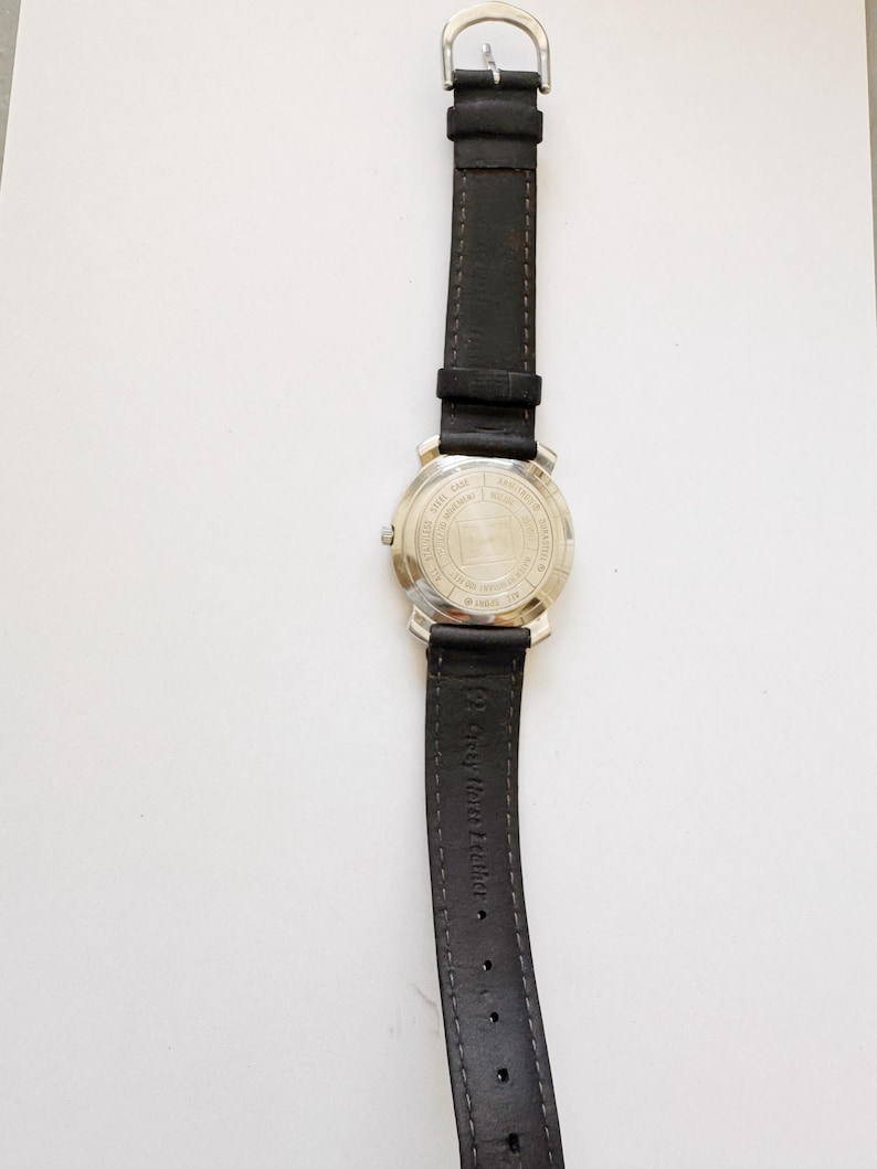 May include: A wristwatch with a black leather band and a silver-colored metal case. The watch band is connected to the case with a silver buckle. The back of the watch case has text and a logo.