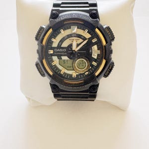 May include: Black and gold Casio digital watch with multiple buttons and a digital display. The watch face has a black background with gold accents and white text. The watch is displayed on a white pillow.