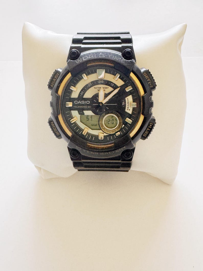 May include: Black and gold Casio digital watch with multiple buttons and a digital display. The watch face has a black background with gold accents and white text. The watch is displayed on a white pillow.
