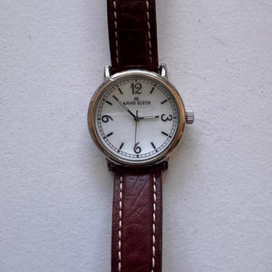 K&ouml;nnte beinhalten: Anne Klein Armbanduhr mit silberfarbenem Geh&auml;use, wei&szlig;em Zifferblatt und braunem Lederarmband. Die Uhr hat schwarze Ziffern bei 12, 3 und 6 Uhr. Das Armband ist mit wei&szlig;em Faden gen&auml;ht.