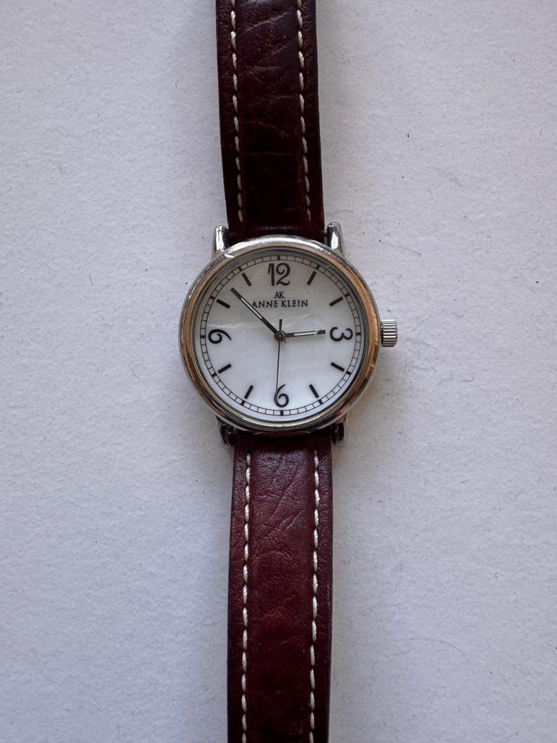 K&ouml;nnte beinhalten: Anne Klein Armbanduhr mit silberfarbenem Geh&auml;use, wei&szlig;em Zifferblatt und braunem Lederarmband. Die Uhr hat schwarze Ziffern bei 12, 3 und 6 Uhr. Das Armband ist mit wei&szlig;em Faden gen&auml;ht.