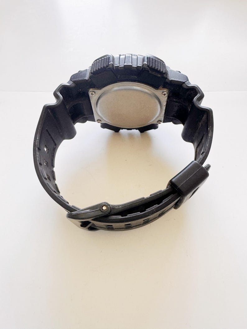 May include: Black digital watch with a textured bezel and band. The watch has a silver-colored back and a black buckle. The watch is designed for sports and outdoor activities.