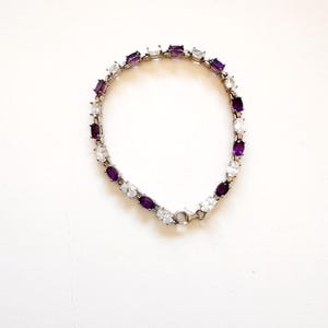 Peut inclure: Un bracelet en argent orné de pierres ovales violettes et transparentes alternées. Le bracelet est posé sur une surface blanche, mettant en valeur son design et le contraste des pierres.