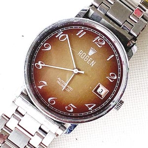 May include: A silver wristwatch with a brown face and silver hands. The watch face has a gradient of brown and features the text "Rogen" and "Automatic Incabloc".