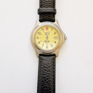 Op de afbeelding: Een Swiss Army quartz horloge met een ronde zilverkleurige metalen kast, een lichtgele wijzerplaat met zwarte Arabische cijfers, een datumvenster op de drie-uurpositie en een gestructureerde zwarte lederen horlogeband met een zilverkleurige gesp.