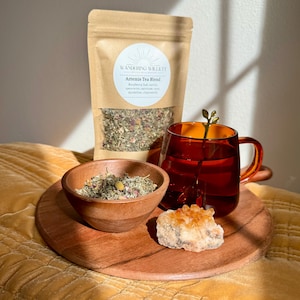 May include: A brown paper bag with a white label that reads "Wandering Willett" and "Artemis Tea Blend". The label lists the ingredients: raspberry leaf, nettle, spearmint, oatstraw, rose, dandelion, chamomile. A wooden bowl filled with loose leaf tea sits next to a red glass mug filled with tea. A small orange crystal sits on a wooden tray.