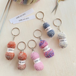 Könnte beinhalten: Sechs Miniatur-Schlüsselanhänger in Form von Wollknäueln in verschiedenen Farben wie Orange, Pink, Blau und Grau. Jeder Anhänger hat ein kleines weißes Stoffetikett mit dem Aufdruck KOMME WAS WOLLE und ist an einem silbernen Metallring befestigt.