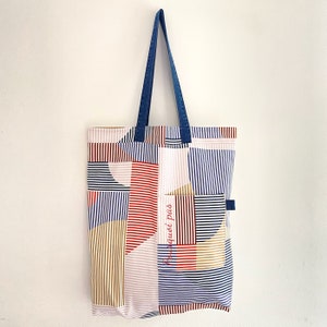 Könnte beinhalten: Eine blaue und weiße gestreifte Einkaufstasche mit einem geometrischen Muster in Rot, Weiß und Braun. Die Tasche hat einen blauen Jeansgriff und der Text "Pas quoi pas" ist auf der Vorderseite gestickt.