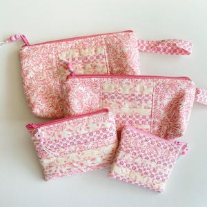 Könnte beinhalten: Vier rosa gesteppte Beutel in verschiedenen Größen. Die Beutel haben ein Blumenmuster und ein Streifendesign. Jeder Beutel hat einen Reißverschluss und einen rosa Reißverschlussanhänger. Der größte Beutel hat einen Griff. Die Beutel liegen auf einer weißen Oberfläche.