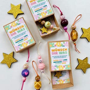 Könnte beinhalten: Mehrere kleine, dekorative Schachteln mit dem Text "Wünsch Dir Was" und Perlenanhängern. Die Schachteln sind geöffnet und zeigen kleine sternförmige Anhänger. Die Anhänger weisen verschiedene Farben und Designs auf, darunter Smiley-Gesichter und Herzen. Goldene Glitzersterne sind um die Schachteln verstreut.
