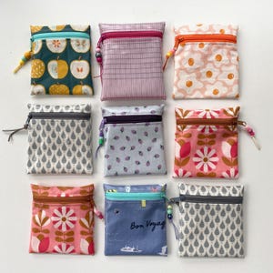 Könnte beinhalten: Eine Sammlung von neun quadratischen Stoffbeuteln mit Reißverschluss. Die Beutel zeigen verschiedene Muster wie Äpfel, Karos, Spiegeleier, Regentropfen, Erdbeeren und florale Designs, jeweils mit einem kleinen Perlenanhänger am Reißverschluss.