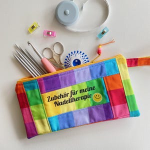 Könnte beinhalten: Ein rechteckiges Stoffetui mit regenbogenfarbenem Karomuster, bedruckt mit dem Text "Zubehör für meine Nadeltherapie" und einem gelben Smiley. Es enthält Stricknadeln aus Metall, eine Häkelnadel, eine Schere und einen Nadeleinfädler.