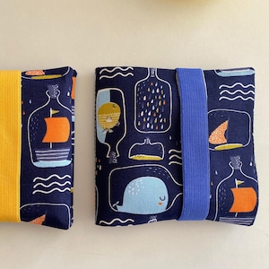 Könnte beinhalten: Zwei blaue Stoffbeutel mit nautischem Thema. Ein Beutel hat einen gelben Riemen und der andere einen blauen Gummizug. Beide Beutel zeigen einen Wal, ein Schiff in einer Flasche und ein Wellenmuster.