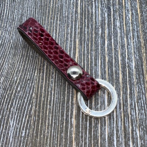 May include: A burgundy leather key fob with a silver-colored split ring. The key fob has a textured, snakeskin-like pattern and a silver-colored rivet. The key ring is attached to the leather strap with a silver-colored metal ring.