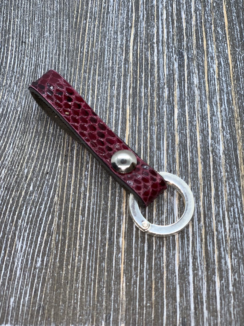 May include: A burgundy leather key fob with a silver-colored split ring. The key fob has a textured, snakeskin-like pattern and a silver-colored rivet. The key ring is attached to the leather strap with a silver-colored metal ring.