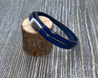 May include: A navy blue leather bracelet with a silver magnetic clasp. The bracelet is wrapped twice around and rests on a small wooden log. The background is a gray wooden surface.