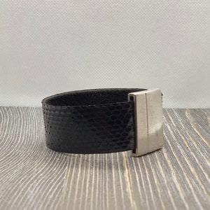 May include: A black leather bracelet with a silver magnetic clasp. The bracelet has a textured, snake-like pattern.