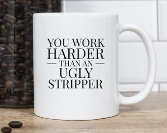 May include: White ceramic mug with the text "YOU WORK HARDER THAN AN UGLY STRIPPER". A glass jar filled with coffee beans is behind the mug. A small white cup is on its side, spilling coffee beans. A vase with greenery is on the left.