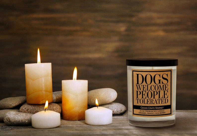 K&ouml;nnte beinhalten: Ein Glas-Kerzengef&auml;&szlig; mit schwarzem Deckel und einem braunen Etikett mit der Aufschrift "Dogs Welcome People Tolerated". Das Etikett enth&auml;lt auch den Text "Cedar Crate Market" und "10 oz Premium Soy Candle Orange Mango Goji Berry". Die Kerze ist von wei&szlig;en und braunen Kerzen sowie grauen Steinen auf einer Holzoberfl&auml;che umgeben.