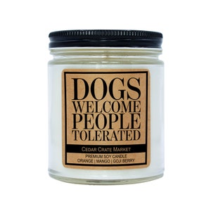 K&ouml;nnte beinhalten: Eine Glas-Kerze in einem Glas mit einem braunen Papieretikett, auf dem "DOGS WELCOME PEOPLE TOLERATED" und "CEDAR CRATE MARKET PREMIUM SOY CANDLE ORANGE | MANGO | GOJI BERRY" steht.