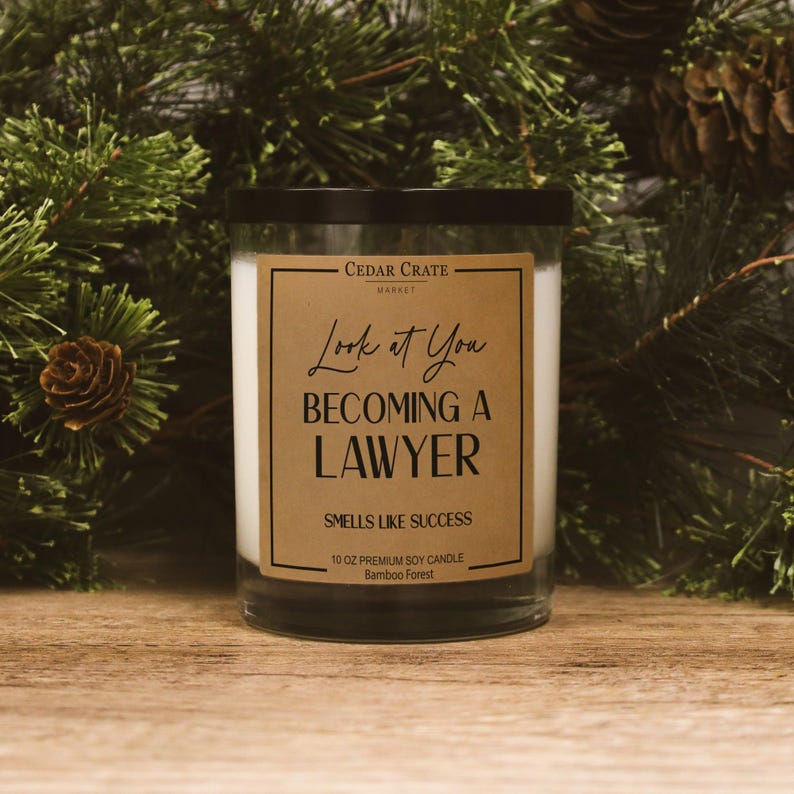 May include: A clear glass jar candle with a black lid and a brown label. The label reads "Look at You Becoming a Lawyer" and "Smells Like Success." The candle is 10 oz and scented with Bamboo Forest. The background features pine branches.