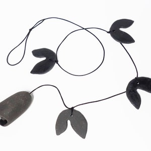 Puede incluir: Un carillón de viento de cerámica negro y gris con cuatro amuletos en forma de hoja y una campana. El carillón está colgado de un cordón negro.