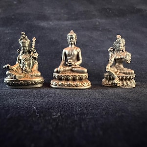 May include: Three small, antique-style Buddha statues in seated positions. The statues are made of a metallic material with intricate details. The central Buddha is in a meditative pose, while the others hold objects. The statues are on a dark surface.