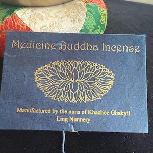 May include: A blue paper bag with gold lettering that reads "Medicine Buddha Incense" and "Manufactured by the nuns of Khachoe Ghakyil Ling Nunnery". A gold lotus flower design is in the center of the bag.