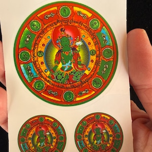May include: A sheet of stickers featuring a detailed, colorful illustration of a deity within a circular design. The central figure is green, surrounded by red, green, and gold accents. Two smaller stickers with the same design are also visible.