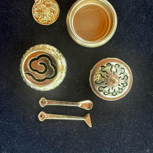 Puede incluir: Un surtido de objetos decorativos de metal dorado, que incluyen un recipiente redondo, tapas con diseños de nubes y flores, y dos piezas pequeñas y alargadas. Los objetos están dispuestos sobre una superficie oscura.