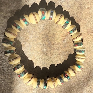 May include: Cream-colored beaded bracelet with decorative accents. The round beads are separated by copper-colored metal rings. Each bead features a band of black, red, and turquoise-colored decorative elements. The bracelet is displayed on a textured, neutral-toned surface.