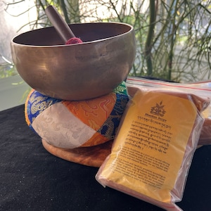 Puede incluir: Un cuenco tibetano de canto con un mazo de madera, un cojín colorido y una bolsa de incienso. La bolsa tiene texto tibetano y las palabras "Shuden Herbs" impresas en ella.