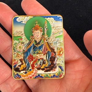 May include: A rectangular, gold-framed religious icon featuring a seated figure with a green halo. The figure wears a crown and colorful robes, holding a staff. The background depicts a landscape with clouds and flowers.