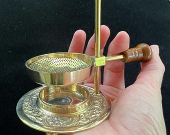 May include: A gold-coloured incense burner with a round, perforated bowl and a decorative base. It features a wooden handle and a vertical post topped with a small sphere. The base has an embossed floral design.